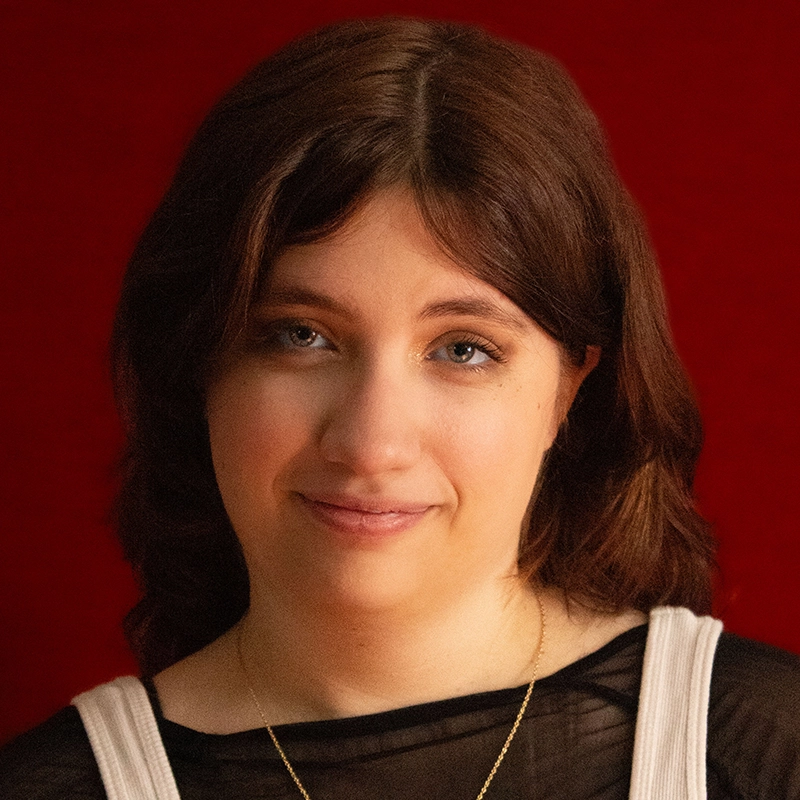 Allison Calabrese, a young woman with medium-length brown hair and blue eyes, smiles gently. She wears a black sheer top with white straps and a gold necklace. The deep red background and warm lighting highlight her facial features.