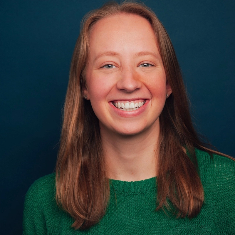 Jade Brathwaite, a young woman with straight, shoulder-length light brown hair, smiles broadly at the camera. She is wearing a green sweater and small stud earrings, posed in front of a solid dark blue background.