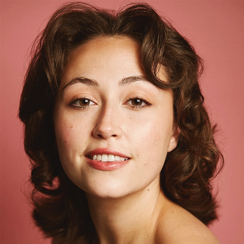 A young woman with wavy brown hair and light skin smiles gently at the camera. She has brown eyes, subtly arched eyebrows, and natural makeup. The background is solid pink, and her shoulders are bare, giving the photo a soft, warm feel.
