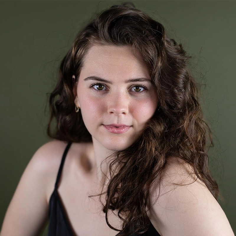 Jade Brathwaite, a young woman with long, wavy brown hair and fair skin, looks directly at the camera. She wears a black spaghetti-strap top and small hoop earring, posing against a plain olive-green background with a hint of a smile.