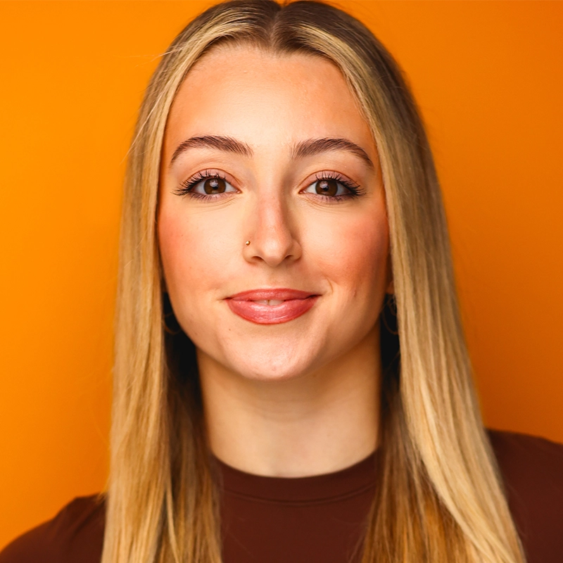 Allison Calabrese, a young woman with long, straight blonde hair and light skin, smiles softly. She has a nose piercing and wears a brown top. The solid bright orange background adds warmth and vibrancy to her portrait.