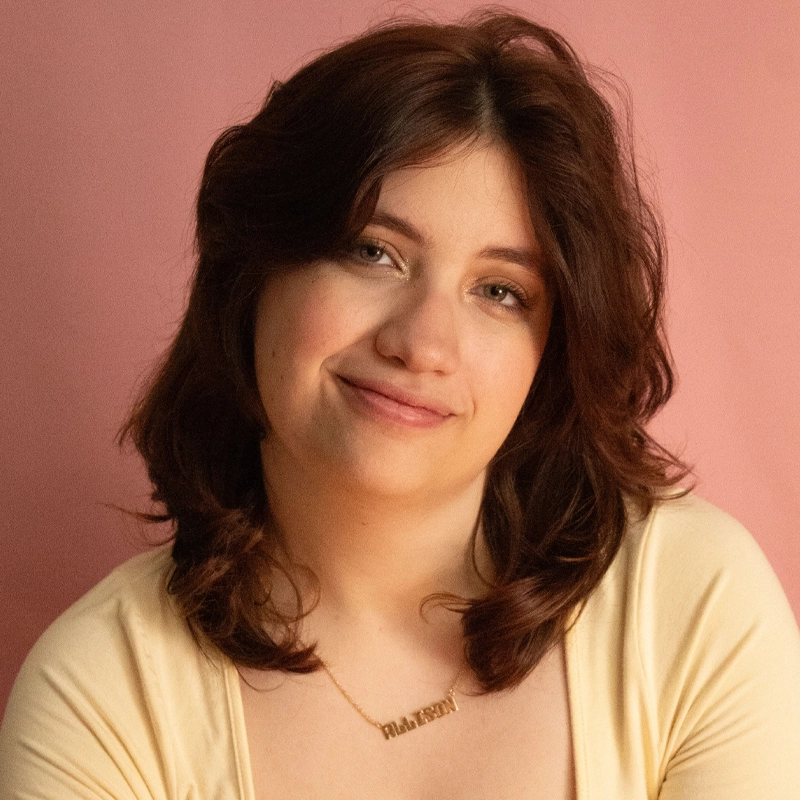 Allison Calabrese, a young woman with shoulder-length brown hair and fair skin, smiles softly. She wears a light yellow top and a gold necklace. The soft pink background creates a warm and gentle atmosphere.