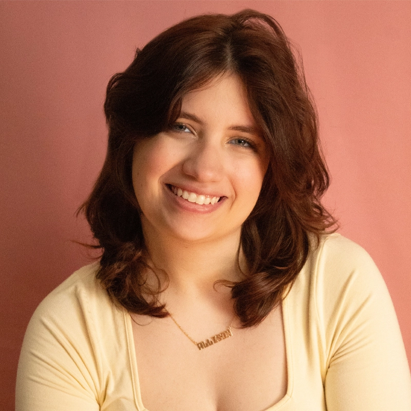 Allison Calabrese, a young woman with shoulder-length brown hair, smiles warmly. She wears a cream-colored top and a gold necklace with small letters. The soft, muted pink background gives the portrait a gentle, inviting feel.