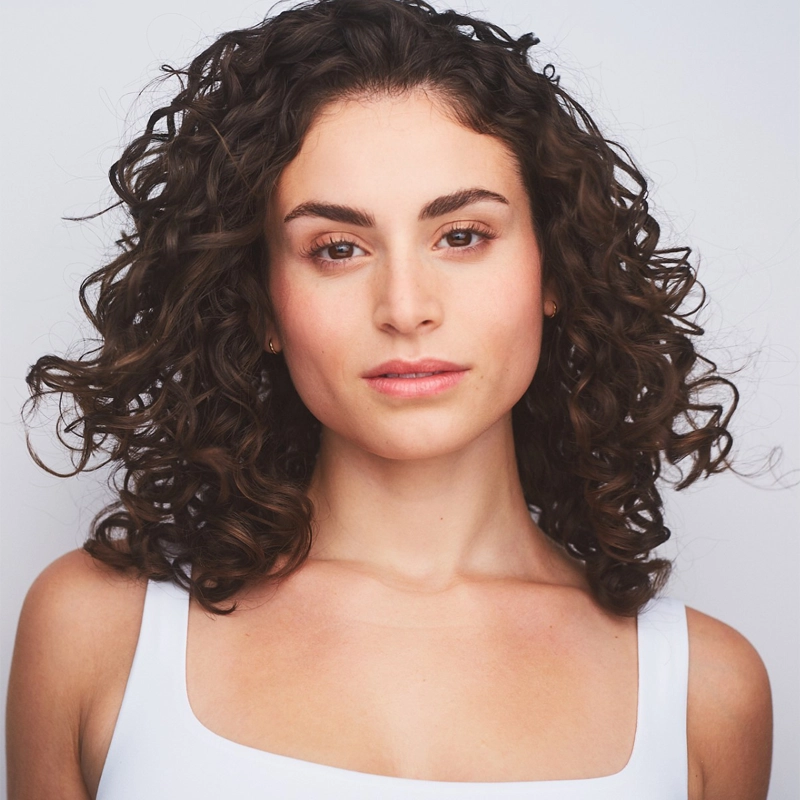 Elana Cantor, a young woman with medium-toned skin and dark, curly hair, gazes confidently at the camera. She has natural makeup, shaped eyebrows, and rosy lips, wearing a white sleeveless top against a light, neutral background.