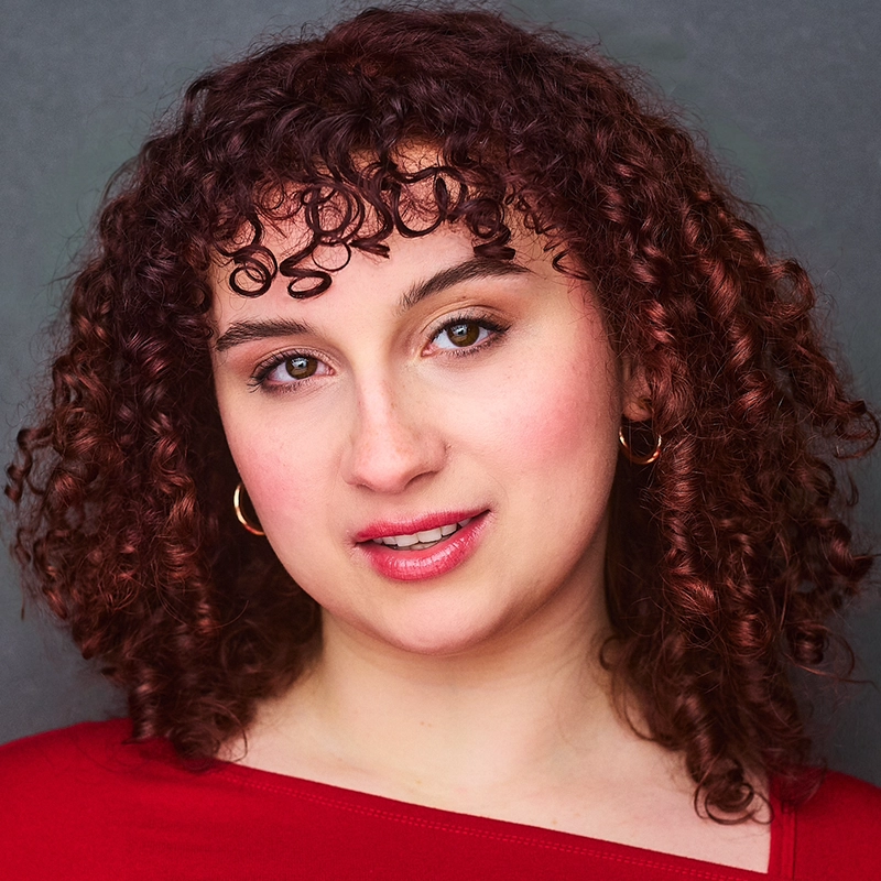 Allison Calabrese, a young woman with curly, dark reddish-brown hair and hoop earrings, looks at the camera with a gentle expression. She wears a red top and light, natural makeup against a plain, neutral gray background.