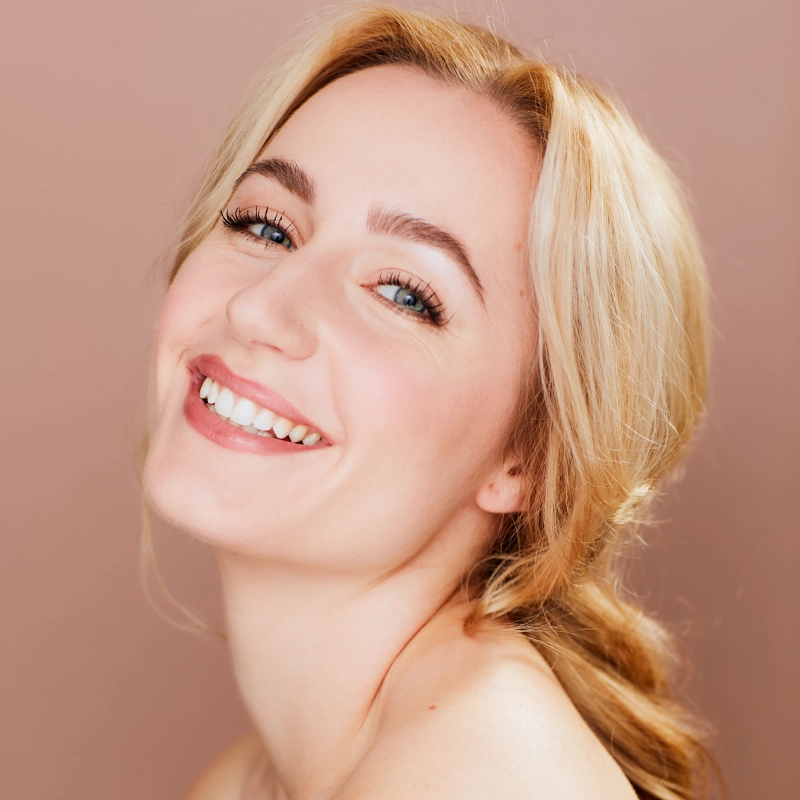 Gabrielle Filloux, a young woman with fair skin and blonde hair, smiles brightly as she looks over her shoulder. She has blue eyes, natural makeup, and poses against a soft pink background, her joyful expression relaxed and genuine.