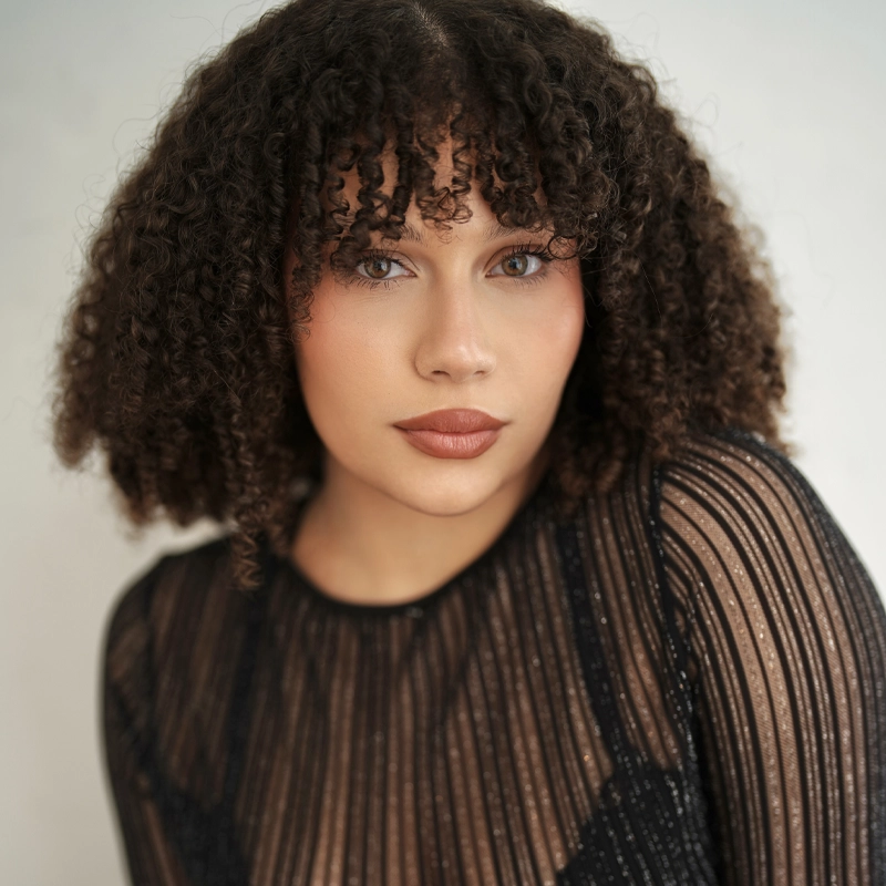 Aaliyana Garcia, with medium skin tone, curly brown hair with bangs, and light eyes, gazes at the camera. She wears a black sheer top with vertical stripes over a black garment. The plain light background highlights her facial features.