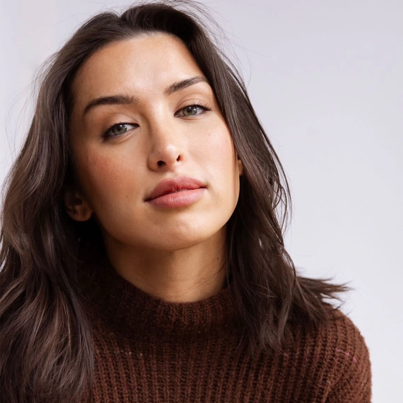 Carissa Gaughran, with long brown hair, light eyes, and natural makeup, wears a chunky brown knit sweater. She looks directly at the camera with a neutral expression against a plain, softly lit background.