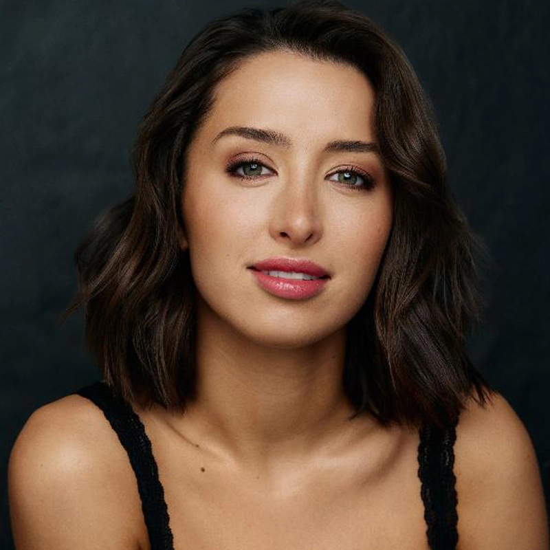 A woman, resembling Carissa Gaughran, with wavy, shoulder-length dark brown hair poses against a dark background. She has light skin, hazel eyes, subtle makeup, and a black sleeveless top with lace straps. She softly smiles at the camera.