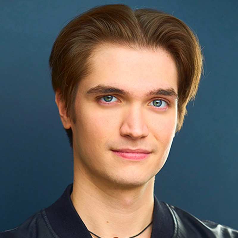 A young man with fair skin, straight light brown hair parted back, and blue eyes, wearing a black jacket and a black necklace, looks at the camera with a slight smile. The photo was taken by Allison Calabrese against a plain dark blue background.