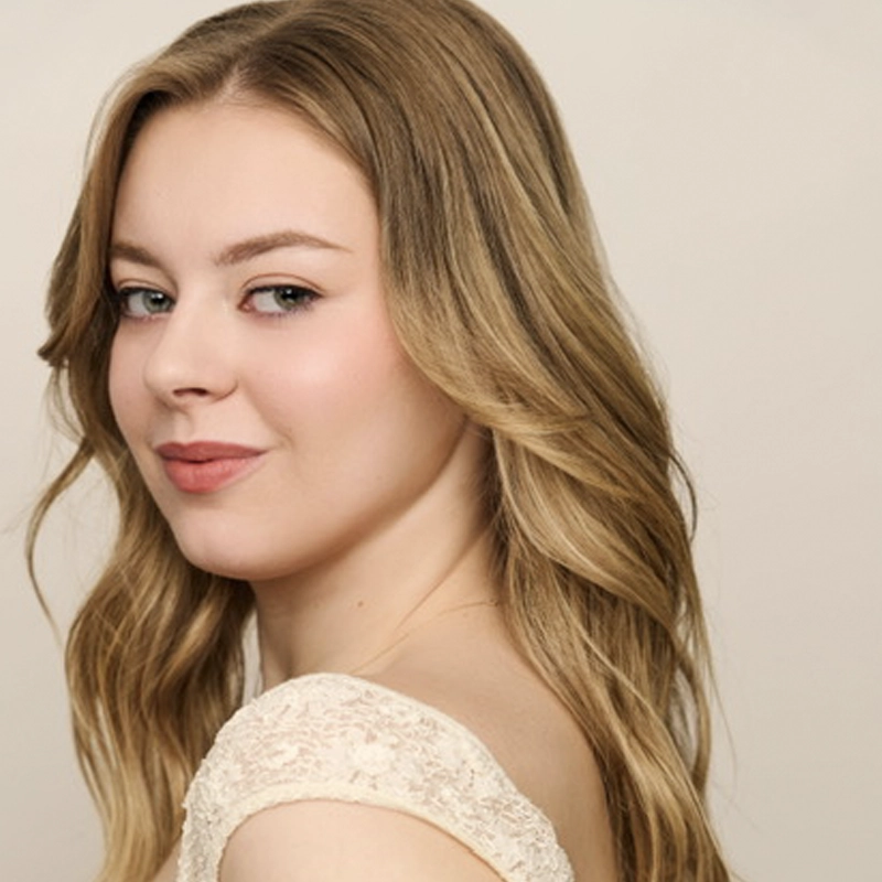 A young woman with long, wavy blonde hair looks over her shoulder at the camera. She is wearing a cream-colored lace top and has a neutral, soft expression against a plain, light beige background.