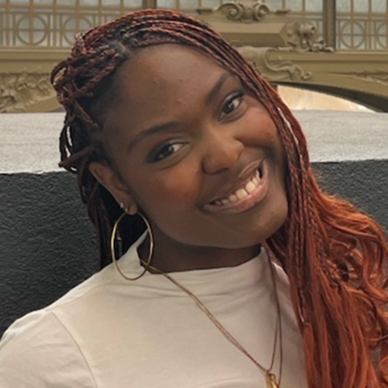 Jade Brathwaite, a young woman with long reddish-brown braids, smiles at the camera. She wears a white top, gold hoop earrings, and a necklace. The background showcases ornate architectural details against a gray wall.