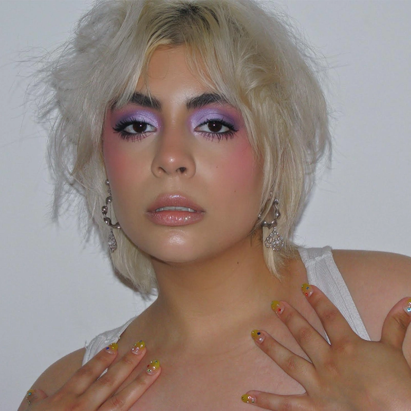 Jade Brathwaite, with short, tousled platinum blonde hair, poses against a plain white background. They wear bold lavender eye shadow, pink blush, glossy lips, dangling earrings, and yellow painted nails near their collarbone.