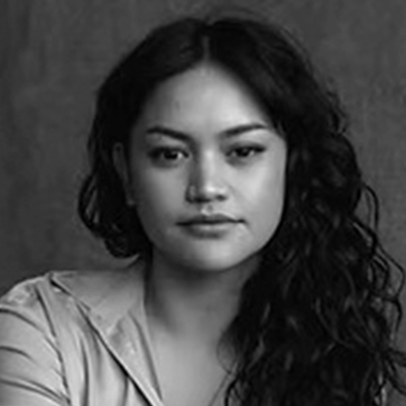 Black and white portrait of a young woman with long, wavy dark hair. In the style of David Socolar, she faces forward with a neutral expression, wearing a light-colored, collared shirt against a softly textured background.