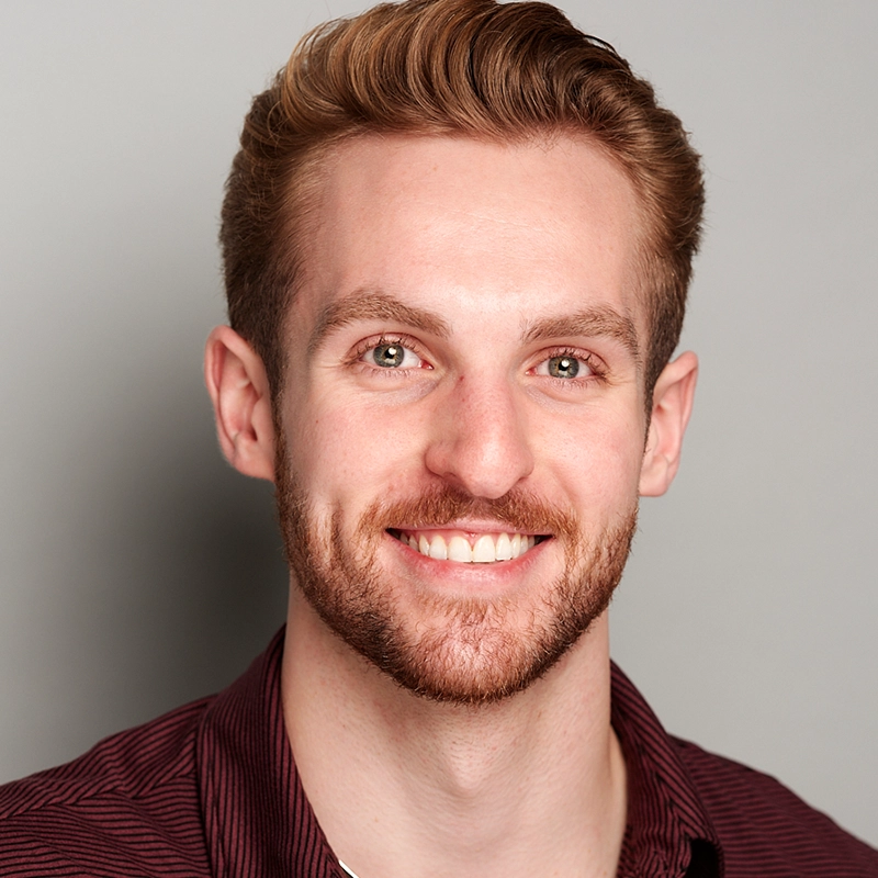 A young man with short, neatly styled light brown hair and a trimmed beard smiles at the camera. Wearing a dark burgundy collared shirt, he poses against a plain light gray background in this Allison Calabrese-inspired portrait.