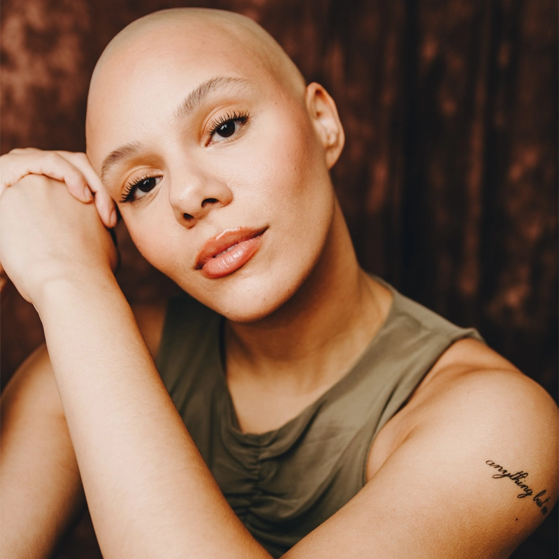A bald person with light skin and brown eyes rests their head on their hand, looking calmly at the camera. They wear a sleeveless olive top and have a small cursive tattoo on their upper arm. The background is brown and softly blurred.