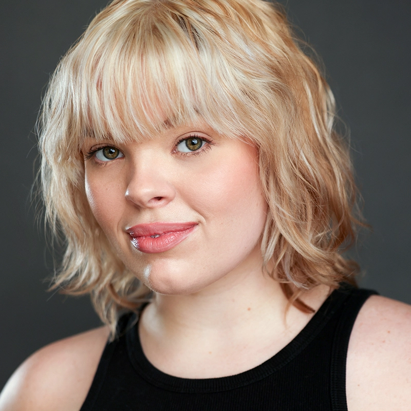 Allison Calabrese, a young woman with short, wavy blonde hair and bangs, smiles slightly. She has fair skin, green eyes, and natural makeup. Dressed in a black sleeveless top, she poses against a neutral gray background.