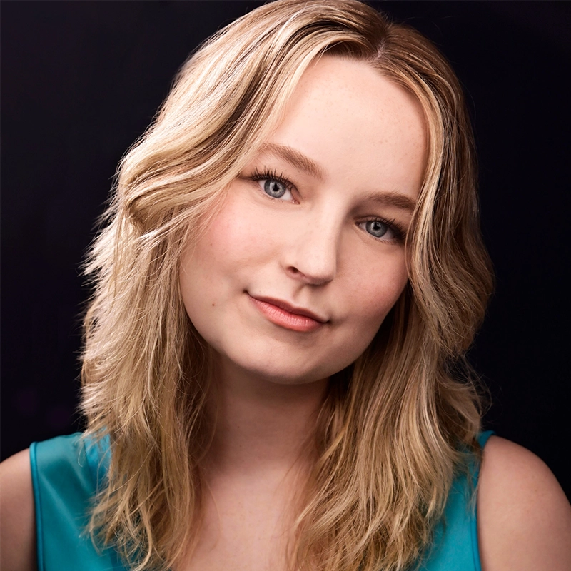 Allison Calabrese, a young woman with fair skin and wavy blonde hair, smiles gently. She wears a teal sleeveless top and natural makeup. The dark background makes her face and hair softly stand out in the light.