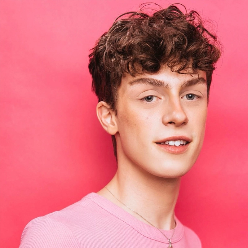 A young person, Jade Brathwaite, with short, curly brown hair smiles softly while facing slightly to the side. They wear a light pink shirt and a thin necklace against a solid bright pink background, giving the image a cheerful, vibrant feel.