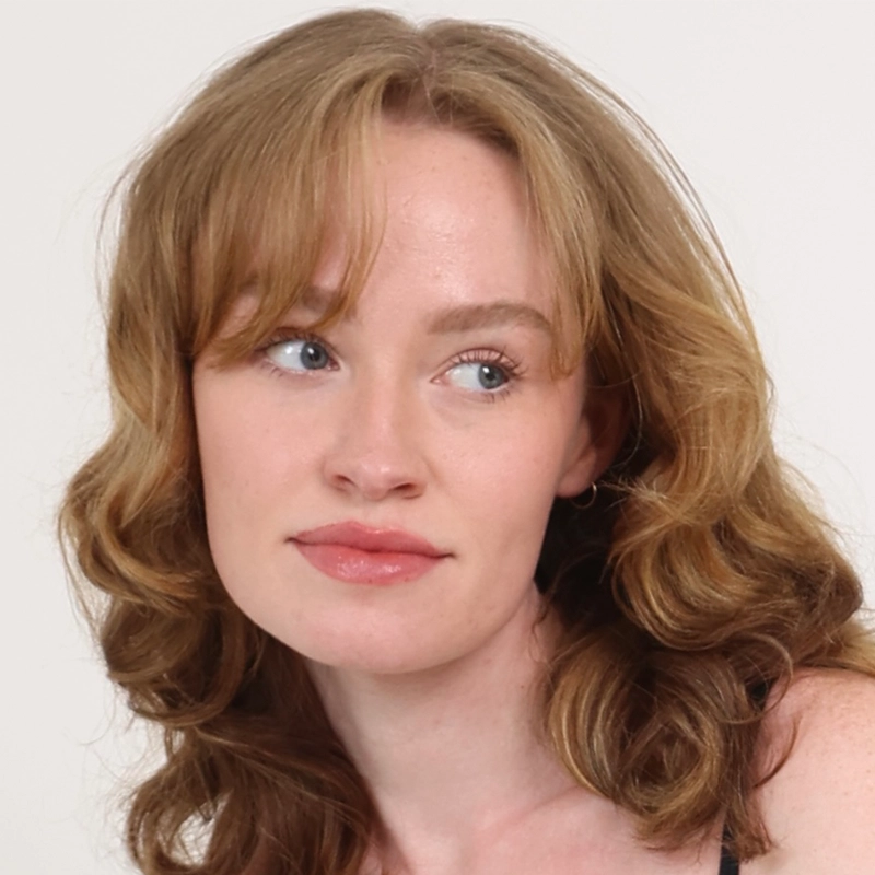 A young woman with wavy, light brown hair and fair skin looks slightly to her left. She has blue eyes, light pink lips, and is wearing a sleeveless top. The background is plain white, and her expression is neutral to thoughtful.