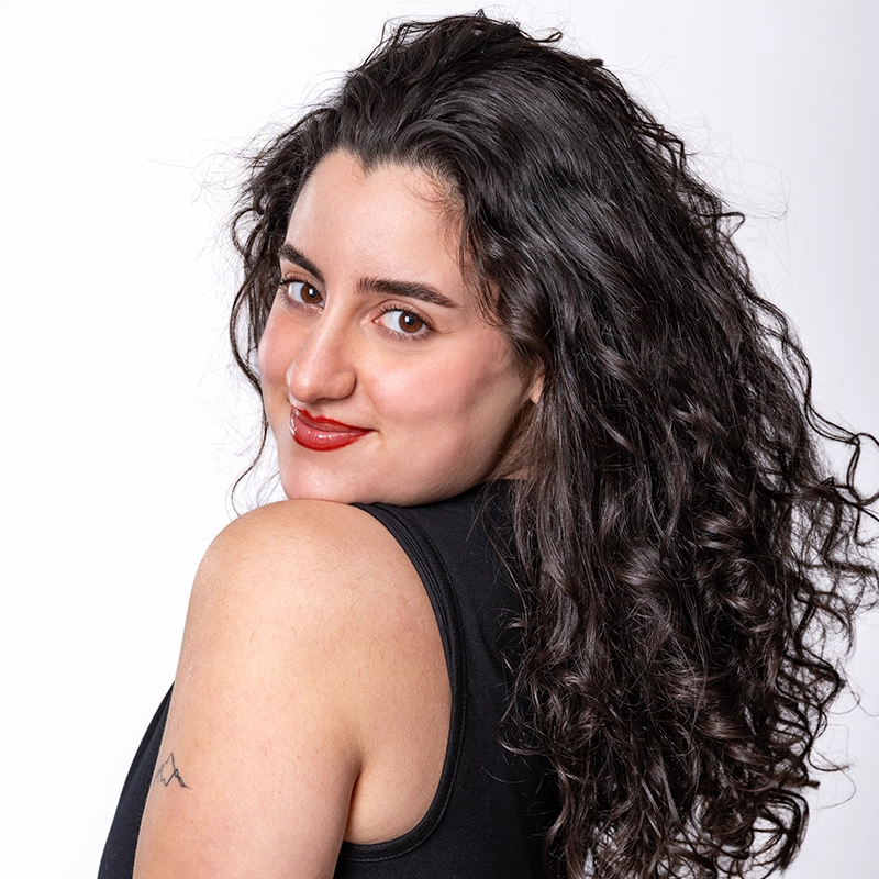 Jade Brathwaite, with long, dark, curly hair, smiles over her shoulder at the camera. She wears a sleeveless black top and red lipstick. Her light skin features a small minimalist tattoo on her upper arm against a plain white background.