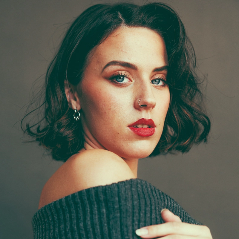 Haiden Pederson, a young woman with fair skin, blue eyes, and wavy dark brown hair, poses against a plain background. She wears red lipstick, a nose ring, and an off-shoulder dark sweater. Her expression is serious as she looks slightly to the side.