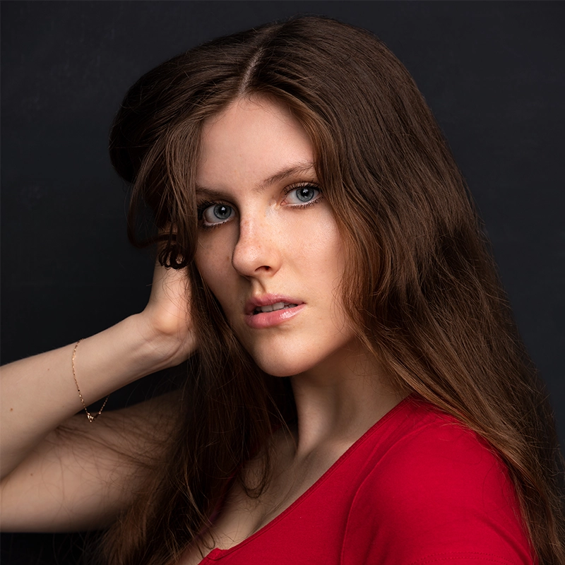 A young woman with long brown hair and blue eyes poses against a dark background. Jade Brathwaite is wearing a red top and a delicate bracelet, her hand gently touching her hair as she gazes directly at the camera with a neutral expression.
