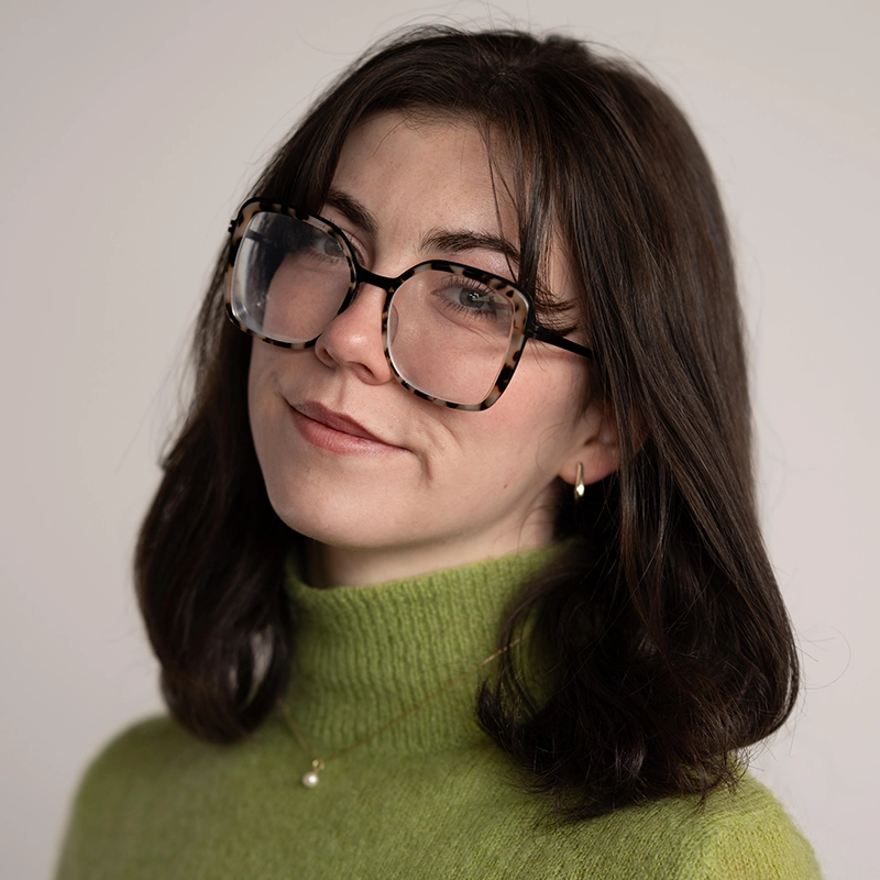 A young woman with shoulder-length dark brown hair wears large, round, tortoiseshell glasses reminiscent of David Socolar’s vintage style, a light green turtleneck sweater, gold hoop earring, and a delicate gold necklace with a single pearl.
