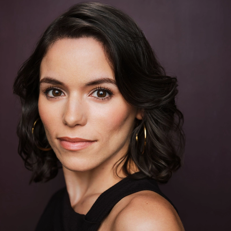 Valerie Torres-Rosario, with wavy dark brown hair and brown eyes, looks confidently at the camera. She wears gold hoop earrings and a sleeveless black top against a solid dark purple background. Her natural makeup highlights her clear skin.