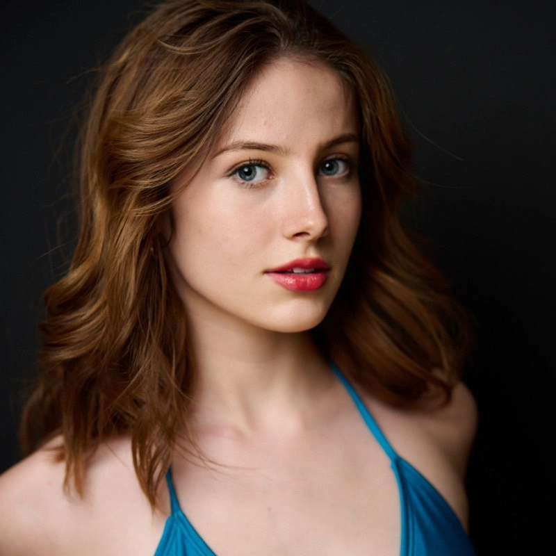 Katherine Shore, a young woman with wavy, shoulder-length brown hair and blue eyes, poses against a dark background. She wears a blue halter top and light, natural makeup with pink lipstick, gazing calmly at the camera.