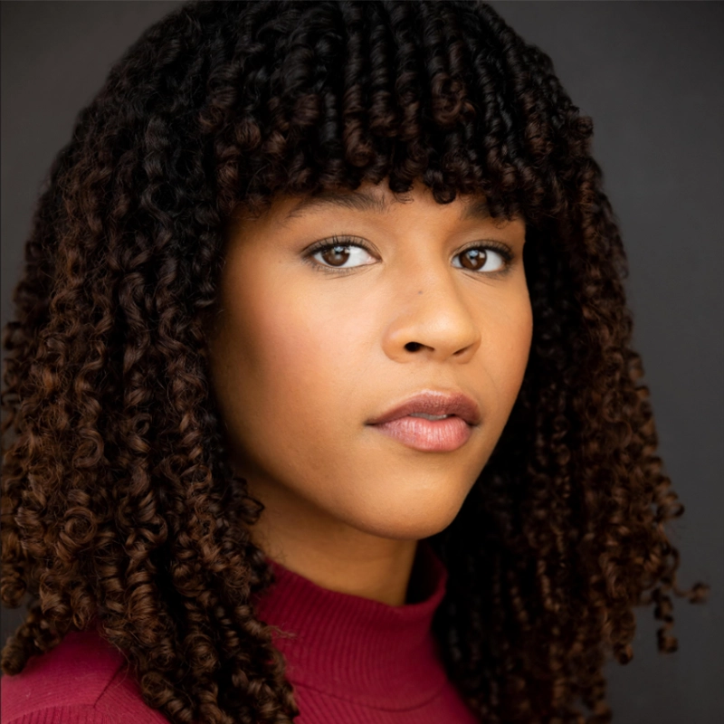 A young woman with medium brown skin and tight, dark brown curly hair, featuring blunt bangs, looks directly at the camera. She wears a high-necked maroon top; the dark background evokes a professional David Socolar studio portrait vibe.