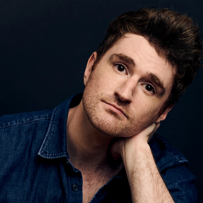 David Socolar, a man with short, wavy brown hair and light skin, wears a blue denim shirt. He gazes at the camera with a neutral expression, resting his head on his left hand. The dark background emphasizes his thoughtful pose.