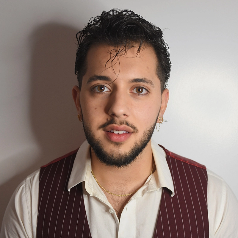 Jade Brathwaite, a young man with short, dark hair and a trimmed beard, stands against a plain white background. He wears a white collared shirt, maroon pinstripe vest, gold hoop earrings, and a thin gold necklace, gazing calmly at the camera.