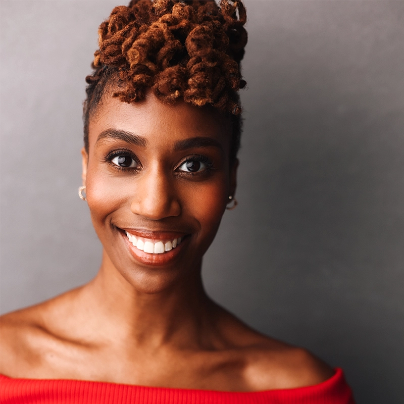A portrait of Sophia Manicone smiling, featuring a woman with dark skin and textured hair styled in an updo, wearing an off-the-shoulder red top.