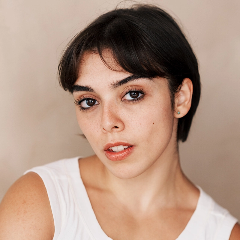 A portrait of performer Sophia Manicone with short dark hair and bangs looking directly at the camera. They have light skin, visible freckles, and are wearing a plain white shirt.
