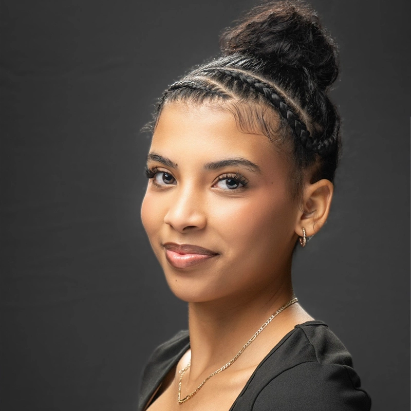 A headshot of Sophia Manicone with dark hair styled in a bun with two braided sections along the front. She is wearing a thin gold chain necklace and small gold hoop earrings.