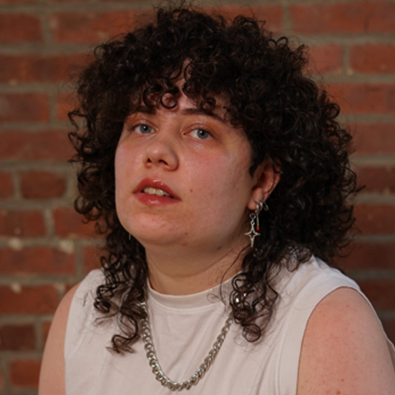 Sofia R. C. Melendez, with curly dark brown hair and fair skin, gazes at the camera in a sleeveless white top, chunky silver chain necklace, and cross-shaped earring. A brick wall serves as the background.