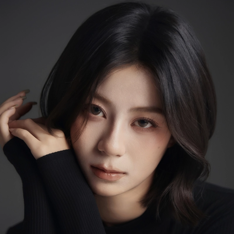 Victoria Chen, a young woman with medium-length black hair and fair skin, gazes softly at the camera. She wears a black top with long sleeves, one hand gently touching her face against a dark gray background for an intimate mood.