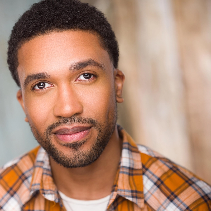 A portrait of performer Sophia Manicone, a man with a short beard looking upward, wearing a plaid button-down shirt.