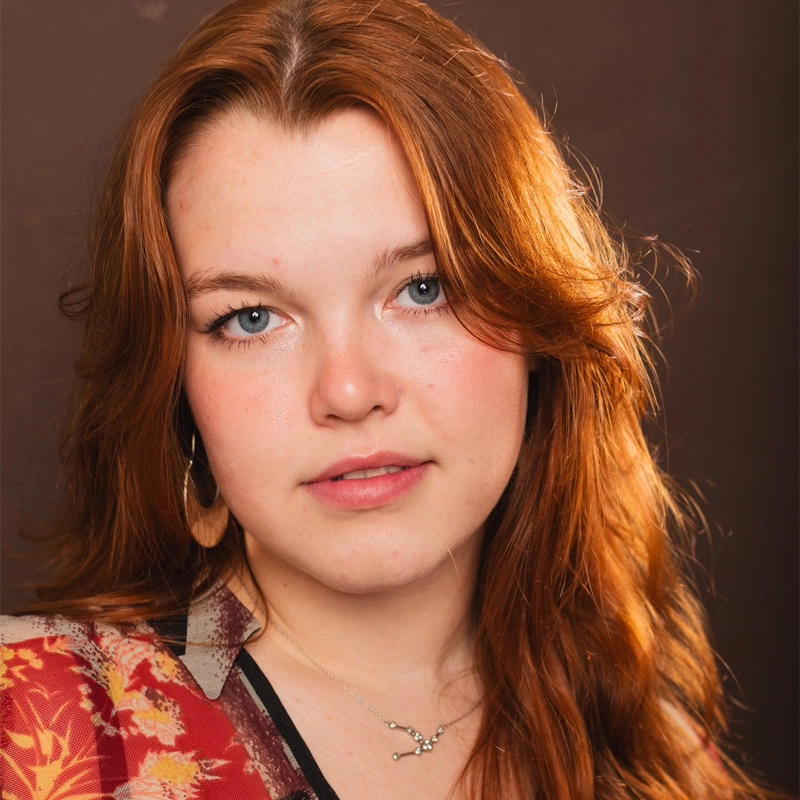 Addison Clover, a young woman with fair skin, long red hair, and blue eyes, looks at the camera. She wears a floral-patterned garment, hoop earrings, and a delicate silver necklace. Warm lighting highlights her features against a soft brown background.