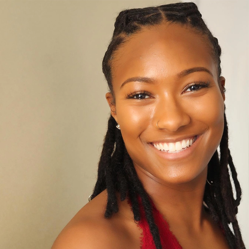 Actress Sophia Manicone smiles, her dark locs pulled back from her face as she wears a red top and a small nose ring.