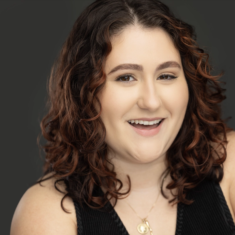 Michaela DeJoseph, a young woman with fair skin and shoulder-length curly brown hair, smiles at the camera. She wears a sleeveless black top and a gold necklace with a round pendant against a smooth, dark gray background.