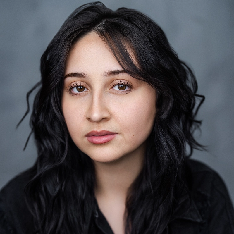 A young woman with long, wavy dark hair and brown eyes looks directly at the camera. She has light skin, natural makeup, and wears a black shirt. The background is a soft gray, creating a calm and neutral setting for this portrait of Gabrielle Driese.