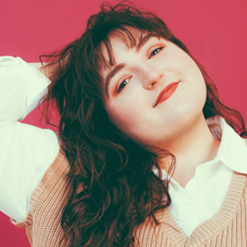 Sofia R. C. Melendez, with wavy dark hair, poses against a pink background, smiling gently. She wears a white collared shirt under a beige knit vest and rests one hand on her head, her makeup featuring red lipstick and subtle eyeshadow.