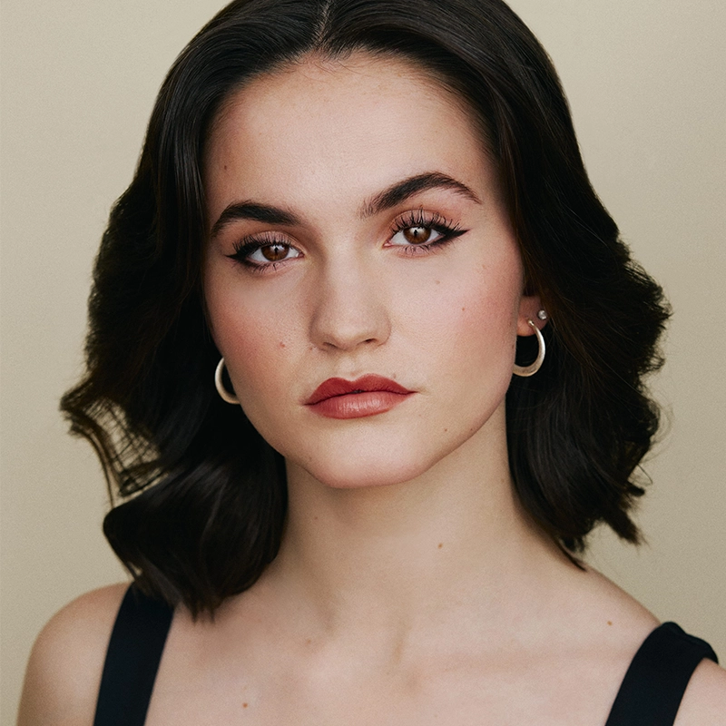 Headshot of performer Sophia Manicone with dark hair and brown eyes looking directly at the camera. She is wearing a black top, small silver hoop earrings, and dark red lipstick.