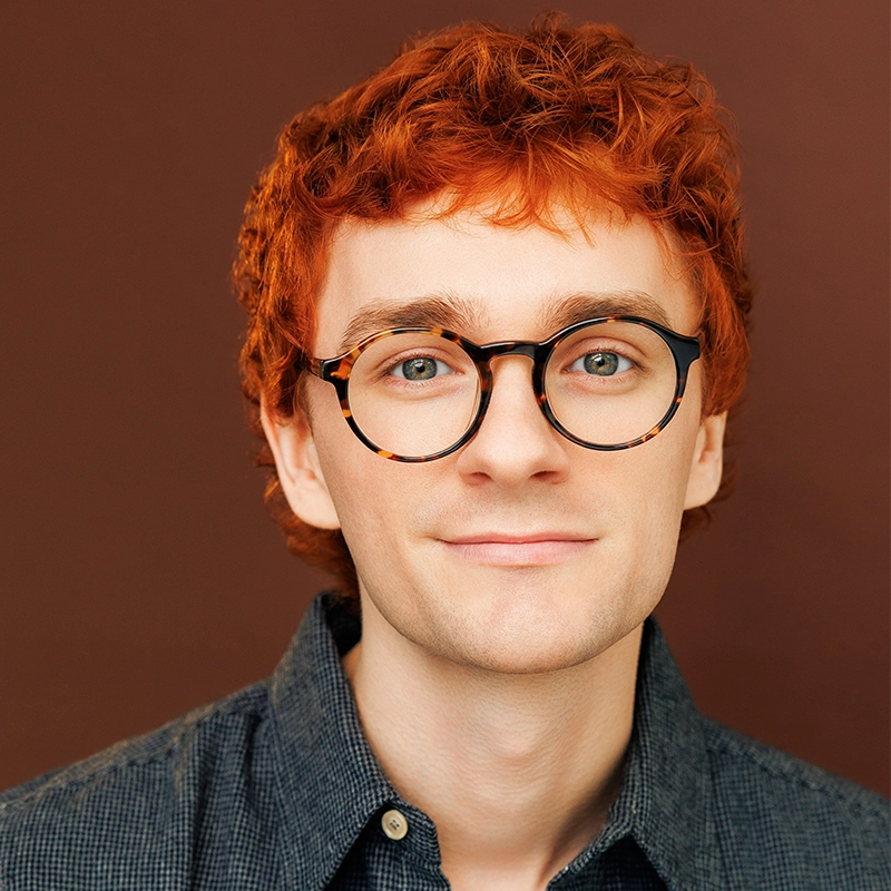 Sophia Manicone, a person with short, curly red hair and round tortoise-shell glasses, looks toward the camera while wearing a dark grey button-up shirt against a solid brown background.