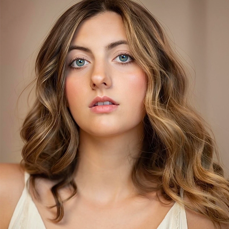 A portrait of actress Sophia Manicone, a young woman with light brown wavy hair and blue-green eyes. She looks directly at the camera with a neutral expression.
