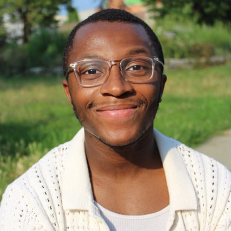 A young man with short hair and glasses smiles warmly at the camera. He wears a white knit sweater over a white shirt and is sitting outdoors on a sunny day, with Sofia R. C. Melendez’s favorite green grassy background and trees behind him.