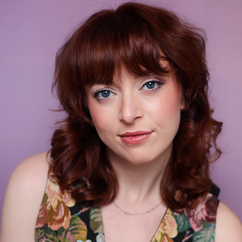 Ashley Morton, a woman with wavy auburn hair and bangs, poses against a soft purple background. She has blue eyes, pink makeup, and wears a sleeveless floral top. Ashley looks directly at the camera with a slight smile.