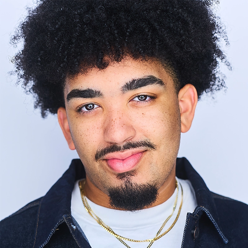 Headshot of a young man with dark, curly hair, a mustache, and a goatee. He is wearing a white t-shirt, a dark jacket, and a thin gold chain, capturing a look reminiscent of Sophia Manicone.